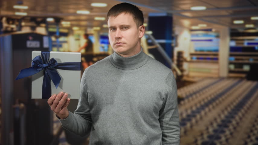 Man in gray turtleneck holding wrapped gift box with navy ribbon, frowning at the present in airport; uncertainty waiting.