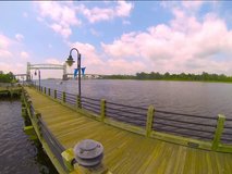 Time lapse video barge moving down Cape Fear River in Wilmington, NC.  Boardwalk in the foreground.  Camera zooms to lift bridge as it drops.  Daytime with clouds moving across the sky.   