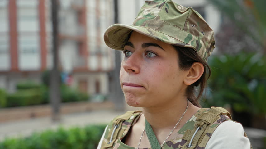 Woman soldier in camouflage cap and tactical vest looks to side, mouth slightly open showing dogtags in building; duty determination.