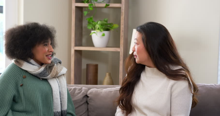 Diverse female friends sitting on couch at home, right woman revealing secret, left holding scarf. Sofa, conversation, duo, surprise, amusement, livingroom, softlight