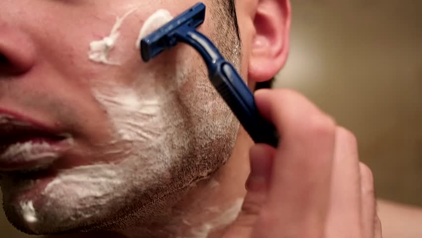 Man shaves his face, that has shaving cream on it, in front of mirror.