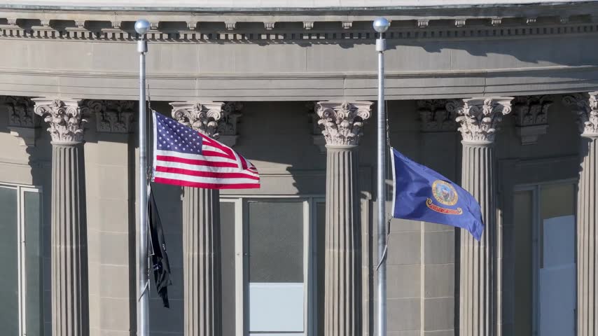 Flags fly at the Idaho State Capital building. American flag and Idaho flag, and the POW flag. Building has classical architecture with columns. Flags wave gently in light breeze