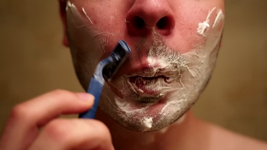 Man shaves his moustache, that has shaving cream on it, in front of mirror.