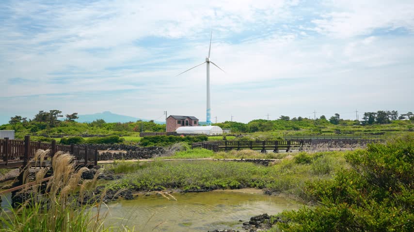 Wooden boardwalk winds through coastal wetlands with wind turbine and pond at Singgyemul Park in Sinchang Jeju South Korea