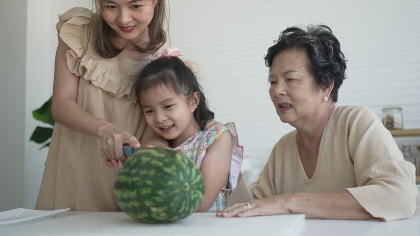 A mother, daughter and grandmother share a healthy eating moment as they cut open a fresh watermelon together in the kitchen.