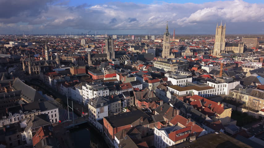 Cinematic aerial Ghent Belgium showcasing medieval towers and historic city center. Drone captures Saint Bavo Cathedral and Belfry spires rising above red rooftops with dramatic clouds.