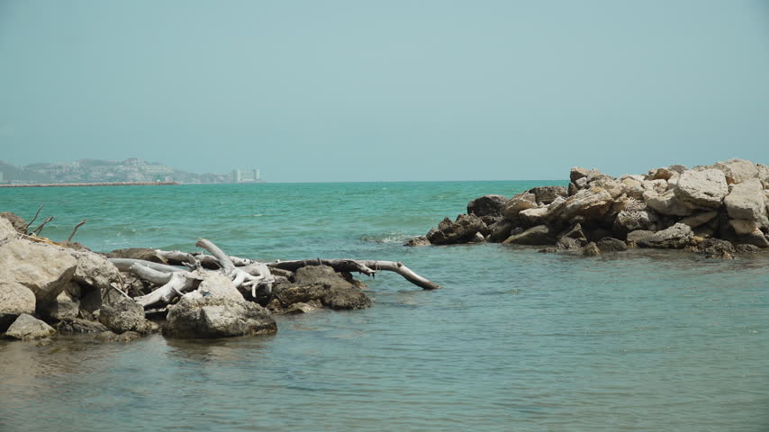 Picturesque sea coastal bay with turquoise water bordered by rocks. In distance of sea bay city appears on hill. Natural stone breakwaters near beach.