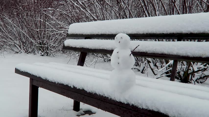 A small snowman sits on a snow-covered park bench surrounded by snow-laden trees in a quiet winter landscape.