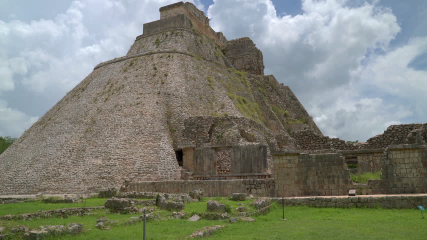 Static low angle wide shot of the iconic oval shaped Pyramid of the Magician at Uxmal Mayan ruins in Yucatan, Mexico under a cloudy blue sky.