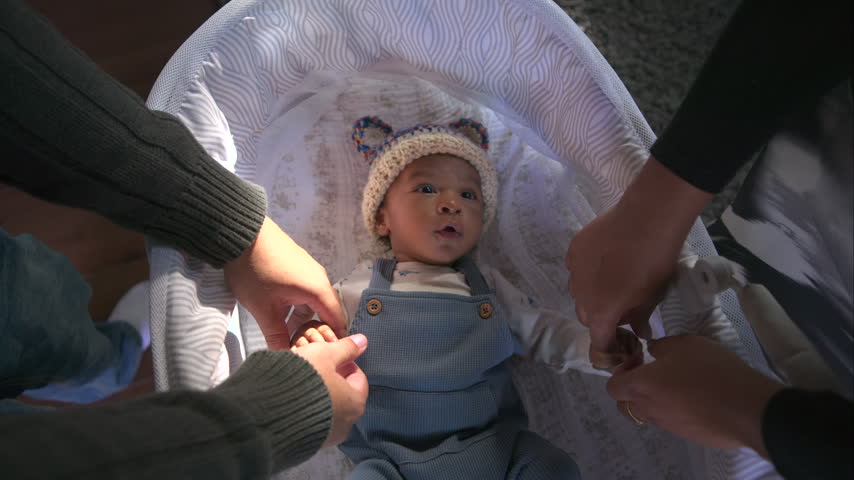 African American young family gently holding tiny hands of their newborn baby lying in white bassinet wearing knitted bear hat and blue overalls at home in soft warm daylight.