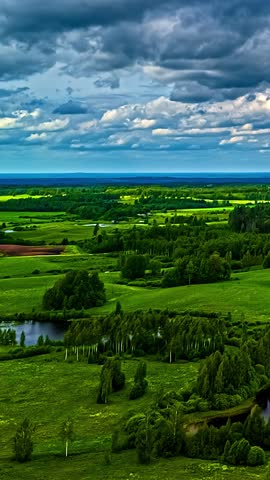Vertical timelapse reveals lush green farmland dotted with small ponds, forest clumps and rolling meadows beneath cloudy sky