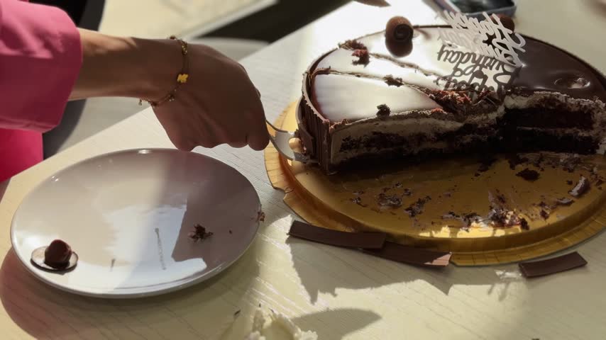 Close-up of hands serving a slice of rich chocolate birthday cake onto a white plate. The action takes place on a bright indoor wooden table illuminated by harsh natural daylight.