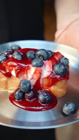 A hand holds a plate with a slice of cheesecake topped with raspberry sauce, sour cream and fresh blueberries. The finished dessert is presented before serving.