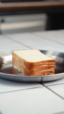 A spoon places sour cream on top of a slice of baked cottage cheese cake on a metal plate. The dessert is being prepared for serving.