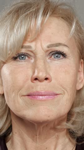 Closeup of a senior woman with blue eyes and blonde hair looking at her face, examining her skin for signs of aging, wrinkles, and crow