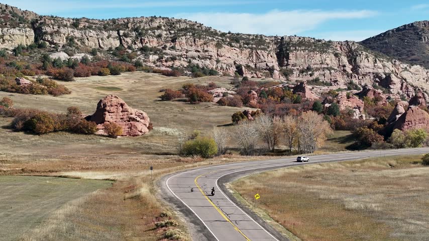 Cinematic 70mm aerial drone shot tracks a solo motorcyclist speeding around a bend near Deer Creek Canyon, surrounded by vivid fall foliage and striking red rock formations in Colorado.