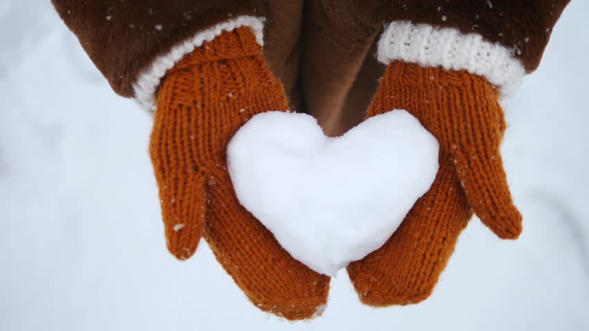 Female hands in warm knitted gloves holding a white heart made of snow during a snowfall, symbolizing love and affection on a cold winter day, perfect for saint valentine