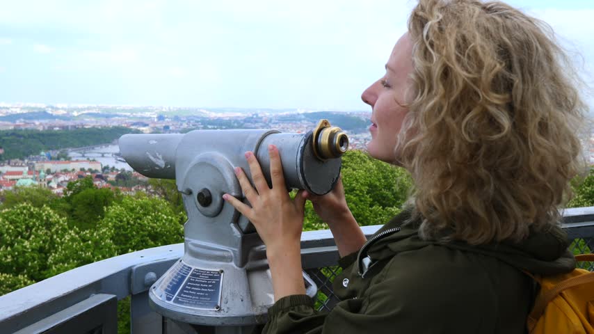 Young blonde tourist with curly hair enjoying the scenic city view from a tower, using a coin operated telescope to observe the panorama of prague, czech republic, from an overlook