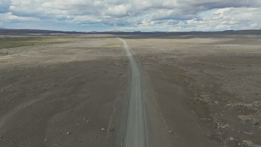 High angle drone shot of a long straight gravel road stretching through a vast, barren volcanic plain in Iceland under a cloudy sky.