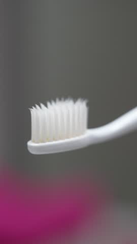 Extreme close-up of a white toothpaste tube dispensing toothpaste onto the bristles of a white toothbrush with a turquoise handle, set against a blurred background.