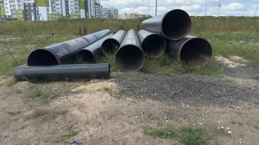 Large metal pipes stacked on ground at construction site with residential buildings in background. infrastructure, development, building, industry, urban.
