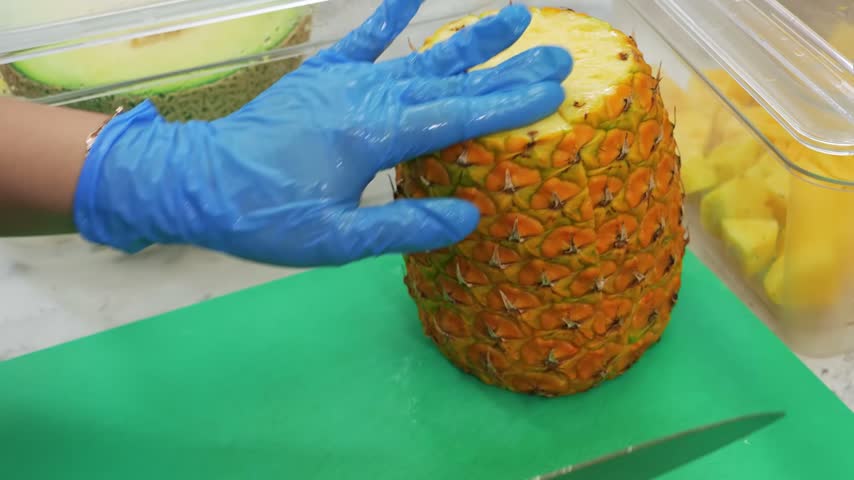 close-up shot of a person wearing blue gloves cutting a fresh, ripe yellow pineapple on a green cutting board in a professional kitchen or food prep area.
