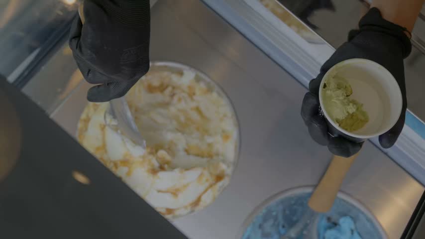 Close-up of artisan gelato being served into a cup by hand, featuring creamy caramel and green flavors in a modern ice cream shop.