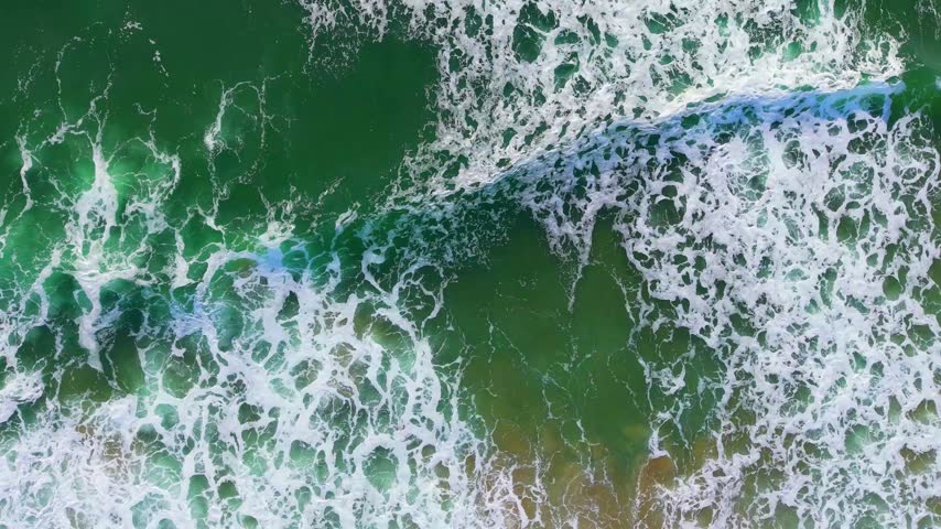 Top-down aerial view of Pacific Ocean waves near Manzanita, Oregon, revealing swirling white surf patterns and deep green coastal water.
