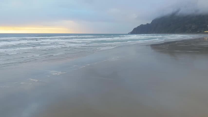 Aerial pan captures a cloudy sunset over Pacific Ocean waves at Manzanita Beach, Oregon, with dramatic clouds and a glowing coastal horizon.
