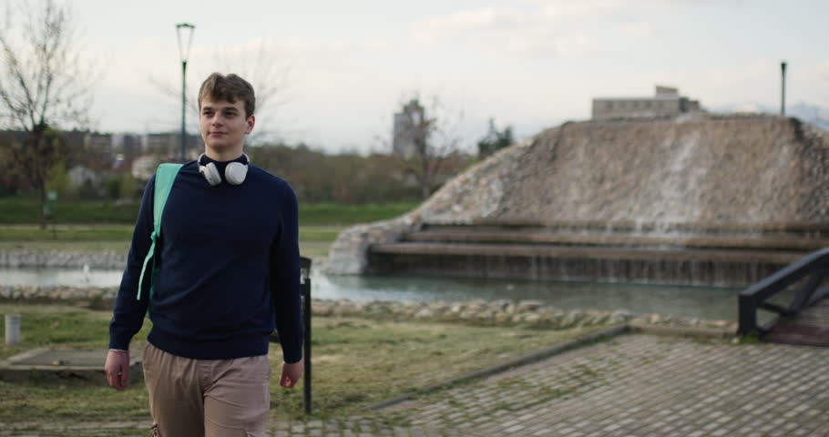A teenage boy with headphones and backpack walks through a park with a fountain and bridge in the background.