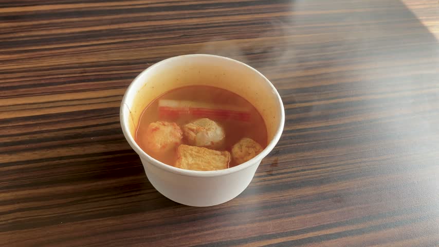 Hot spicy tom yum soup served in a white paper bowl with fish cakes, crab stick, and meatballs on a wooden table.