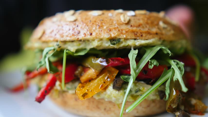 Close up view of a freshly made vegan bagel sandwich filled with creamy hummus, roasted red and yellow peppers, and fresh arugula, showing the detailed texture of the wholesome ingredients