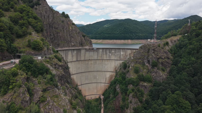 Elevated footage reveals the curved dam beneath rugged slopes while the reservoir extends into Romanian mountains.