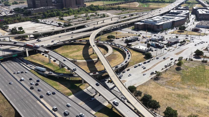 Directly overhead, cars queue along a looping freeway ramp that curls over dry grass and a commercial strip of restaurants and small businesses. The geometry of the concrete loops and the shadows they throw dominate this Dallas, Texas aerial.