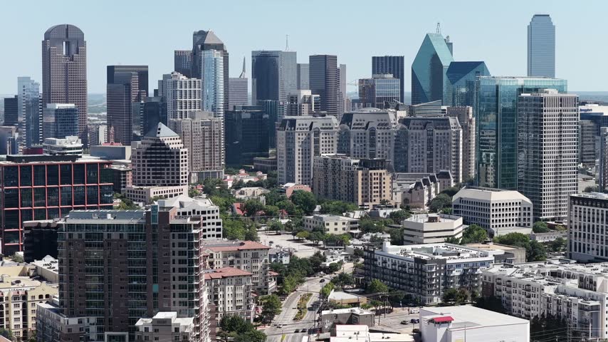 Brick and stucco mid-rise buildings of Uptown fill the foreground while the whole downtown Dallas skyline stands behind them in clear Texas air. A construction crane swings above the district and shaded streets run between the blocks.