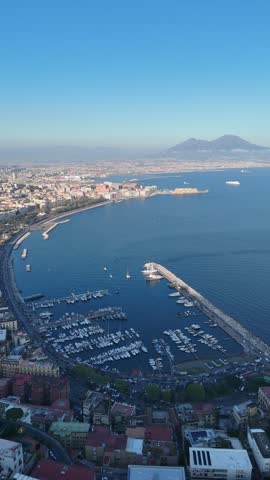 An aerial view of the Italian city of Naples showing the sea, coastline, ships, boats, port, and numerous buildings.