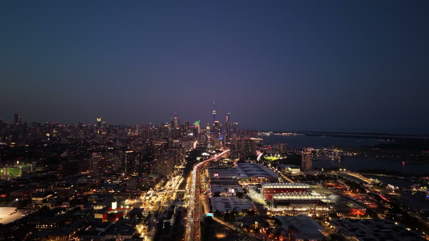 Elevated dusk skyline over downtown Toronto with glowing tower lights, busy roadway leading toward city core, dark waterfront, and expansive blue evening sky above layered high-rises