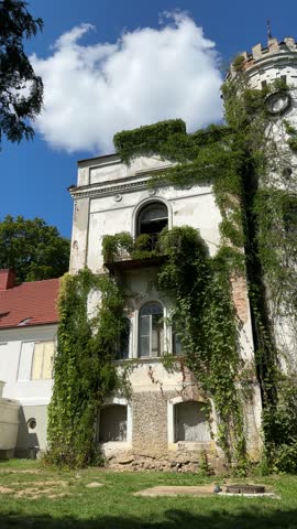 An abandoned old manor house with a white round tower and overgrown ivy under a blue sky with fluffy clouds.