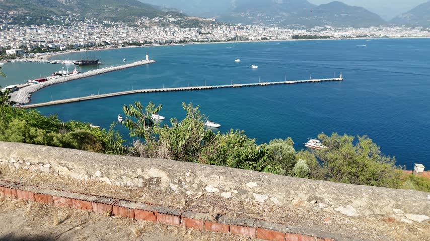 A breathtaking panoramic aerial video capturing the historic Red Tower (Kızıl Kule), the bustling harbor of Alanya, and the Mediterranean cityscape. Numerous tourist pirate ships, yachts, and motorboats cruise across the sparkling blue sea on a sunny summer day.