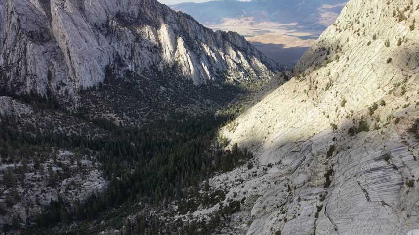 Aerial flight above a steep granite canyon reveals Whitney Portal Road winding through conifer forest toward sunlit Owens Valley in California