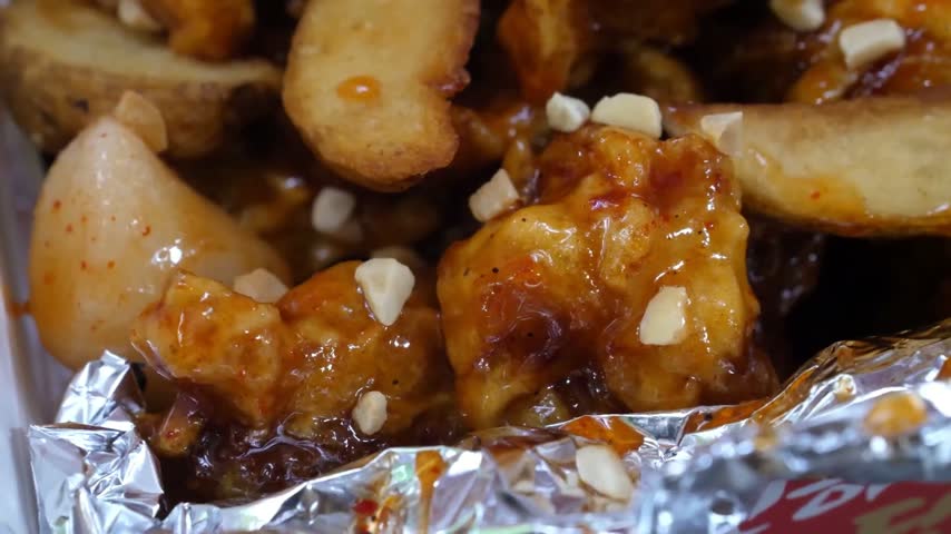 A macro, slow-motion shot of crispy fried chicken pieces coated in a glossy honey garlic glaze, served with golden potato wedges and garnished with crushed peanuts on aluminum foil.
