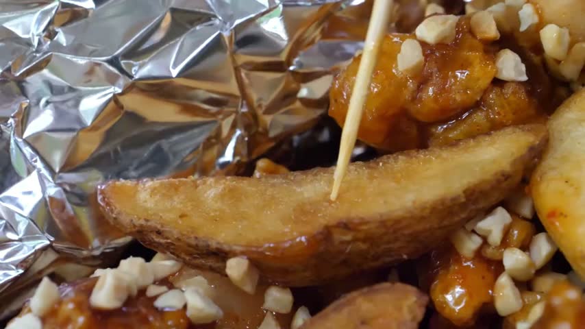 close-up, macro shot captures a golden-brown, crispy potato wedge being pierced by a small wooden skewer or toothpick. The potato wedge is held over a bed of savory, glossy glazed chicken pieces coate

