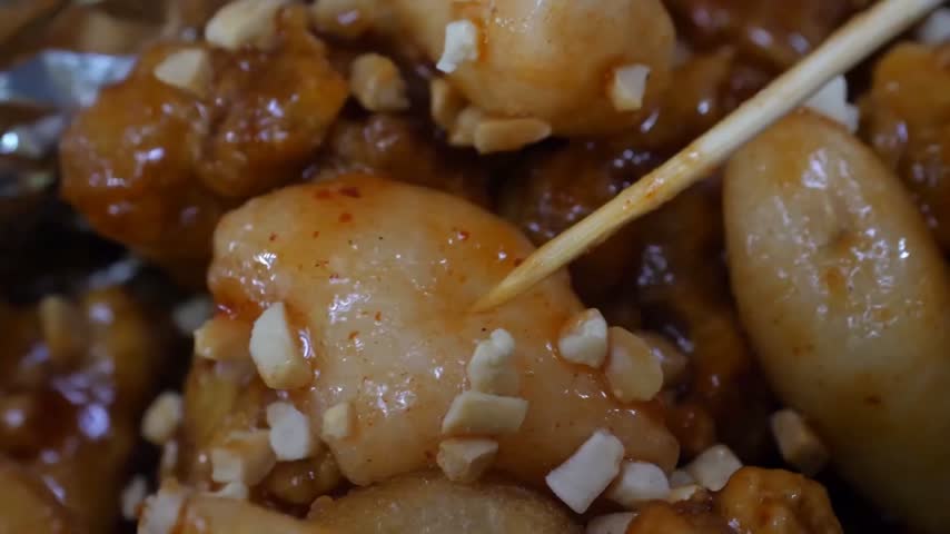 An extreme close-up, high-utility macro shot of popular Korean street food. The footage captures a wooden skewer lifting a piece of chewy Korean rice cake (tteokbokki) and crispy fried chicken (dakgan
