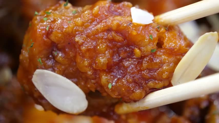 An extreme close-up macro shot of wooden chopsticks lifting a piece of crunchy Korean fried chicken. The succulent boneless chicken is perfectly fried, thickly coated in a glossy, vibrant orange-red 