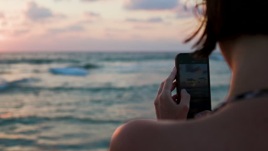 A woman is standing on the beach and photographing the bright sunset of the sea. A girl holds her phone in her hand and takes pictures of the content. Sunset on the beach.
