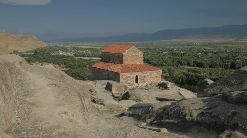 Static handheld shot of an old, abandoned brick church situated on the rocky terrain of the ancient cave city Uplistsikhe, Georgia. A scenic green valley and distant mountains are visible in the background under a clear sky.