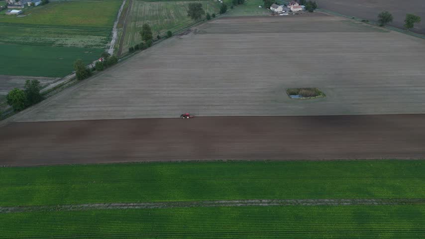 Aerial footage over a broad cultivated field with a single round hay bale, green crop strips, hedgerows, and patchwork farmland extending toward a rural horizon, illustrating harvest season and agricultural land use.