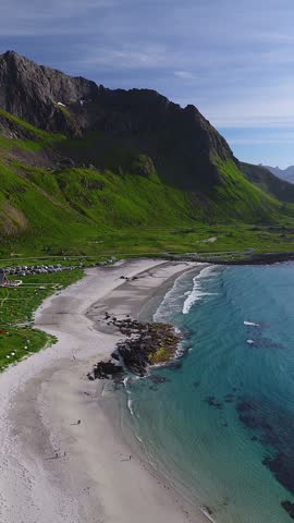 Aerial drone shot reveals the sandy shores, crashing waves, and towering mountain peaks around Skagsanden beach in Flakstad, Lofoten, Norway.