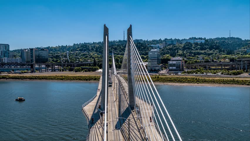 Aerial drone flyover approaching Tilikum Crossing bridge spanning the Willamette River in Portland, Oregon, with the modern cable stayed bridge, Portland skyline and waterfront visible on a clear summer day.