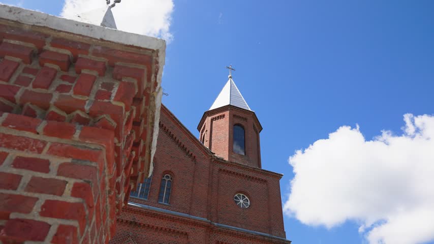 Striking low angle footage pans upward, revealing red brick church towers under a vivid blue sky with scattered clouds. Sunlight highlights the architectural detail in this steady, wide angle shot.